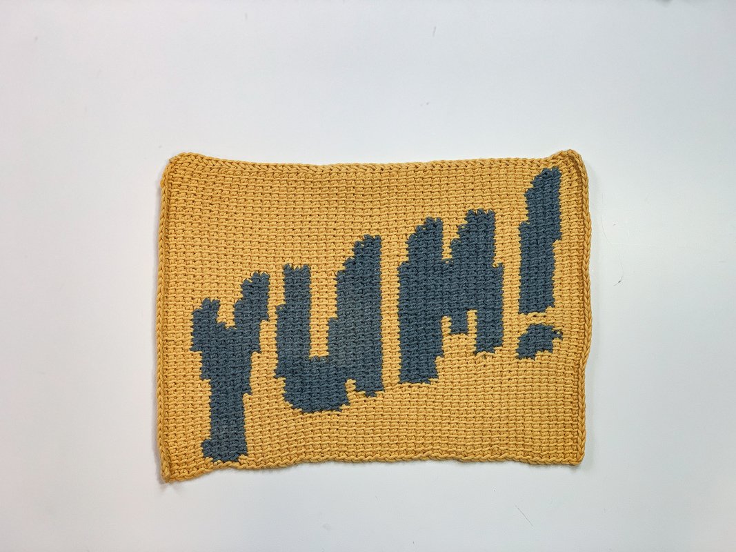 Zweifarbige Häkelarbeit mit 'YUM!'-Motiv und sichtbarer Maschenstruktur