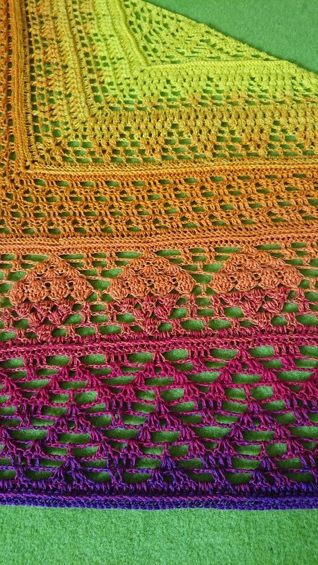Nahaufnahme eines gehäkelten Dreieckstuchs mit Farbverlauf von Gelb bis Violett und abwechslungsreichen Loch- und Rautenmustern.