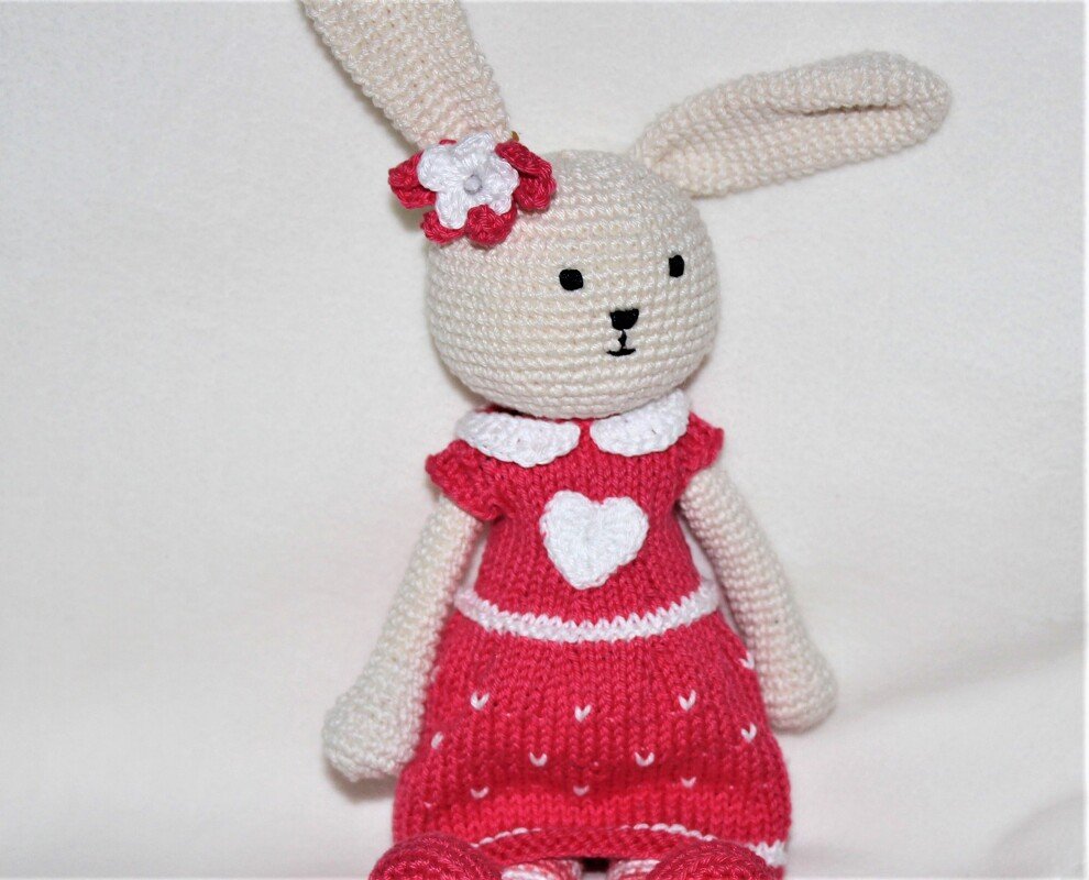 Gehäkelter Amigurumi-Hase mit himbeerrotem Kleid, weißem Herz und Blume am Ohr