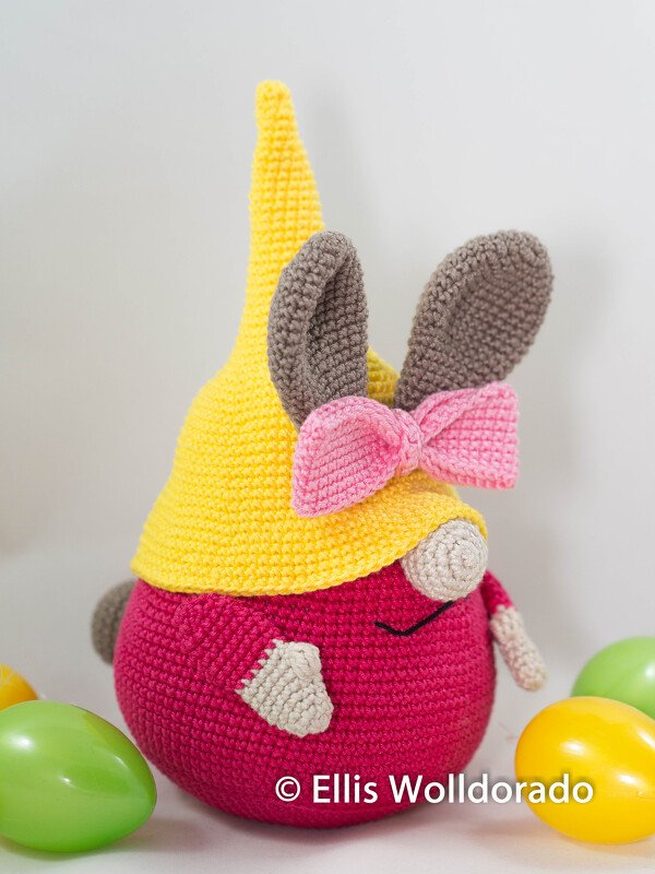 Gehäkelter Amigurumi-Gnom in Pink und Gelb mit Hasenohren und Schleife, umgeben von Ostereiern