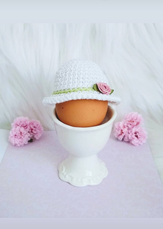 Braunes Ei im weißen Eierbecher mit weißem gehäkeltem Hut, grünem Band und rosa Rose