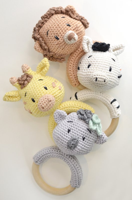 Vier gehäkelte Baby-Greifringe in Form von Löwe, Zebra, Giraffe und Nashorn mit Holzringen auf hellem Hintergrund.