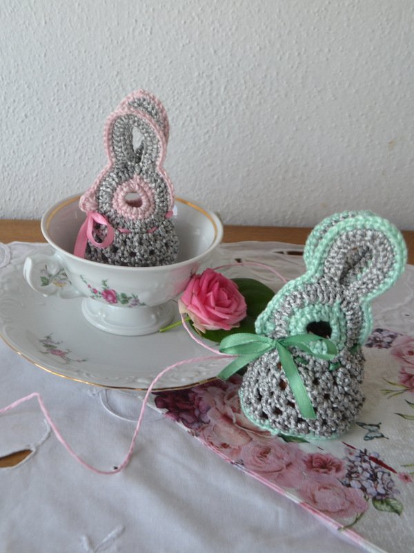 Kompakter gehäkelter Hase mit offenen Maschen im Körper, gehäkelten Ohren und grüner Satinschleife; im Hintergrund ein zweiter Hase mit rosa Rand.