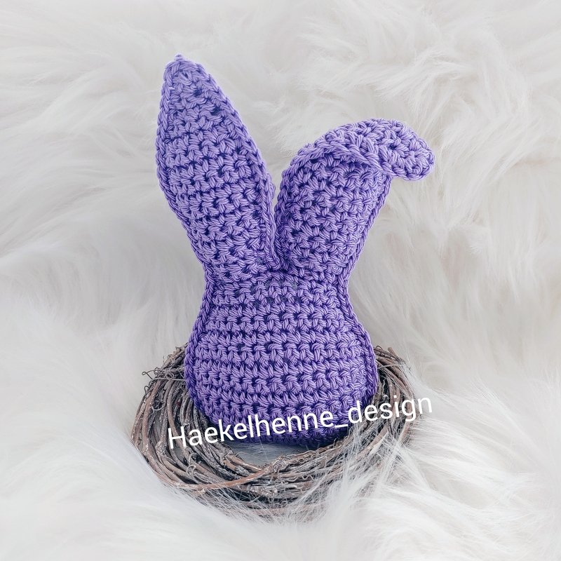 Gehäkelter lila Osterhase mit langen Ohren im kleinen Zweignest auf weißem, flauschigem Hintergrund.