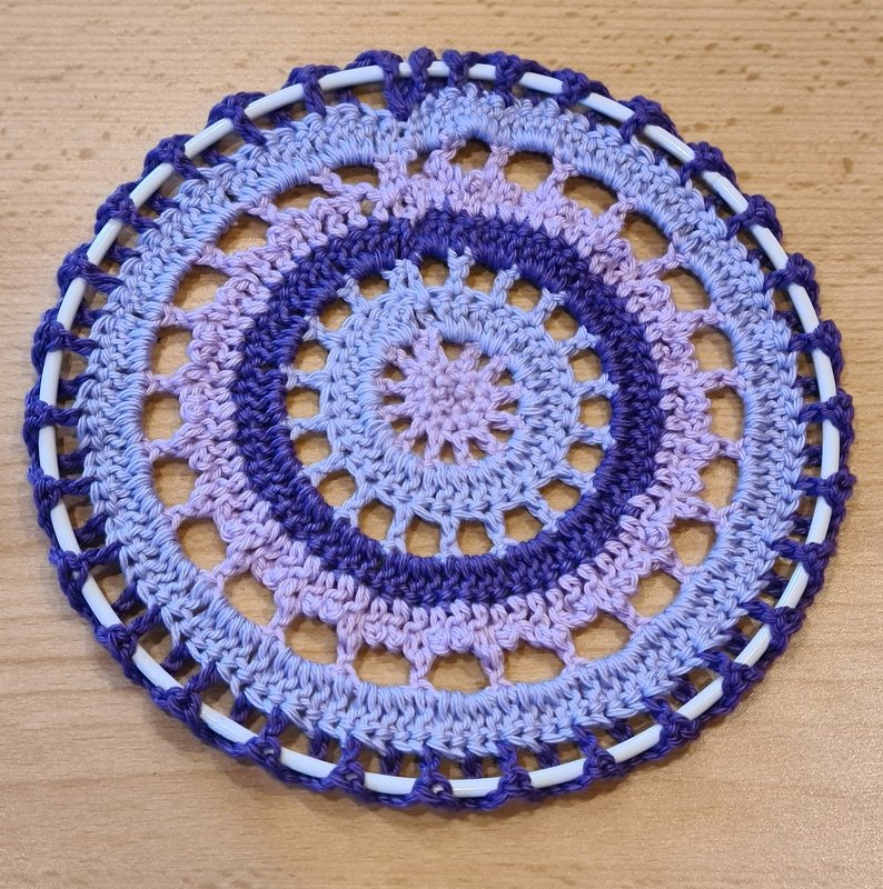 Nahaufnahme eines runden gehäkelten Mandalas in Lila, Lavendel und Hellblau um einen weißen Ring auf einer Holzoberfläche.