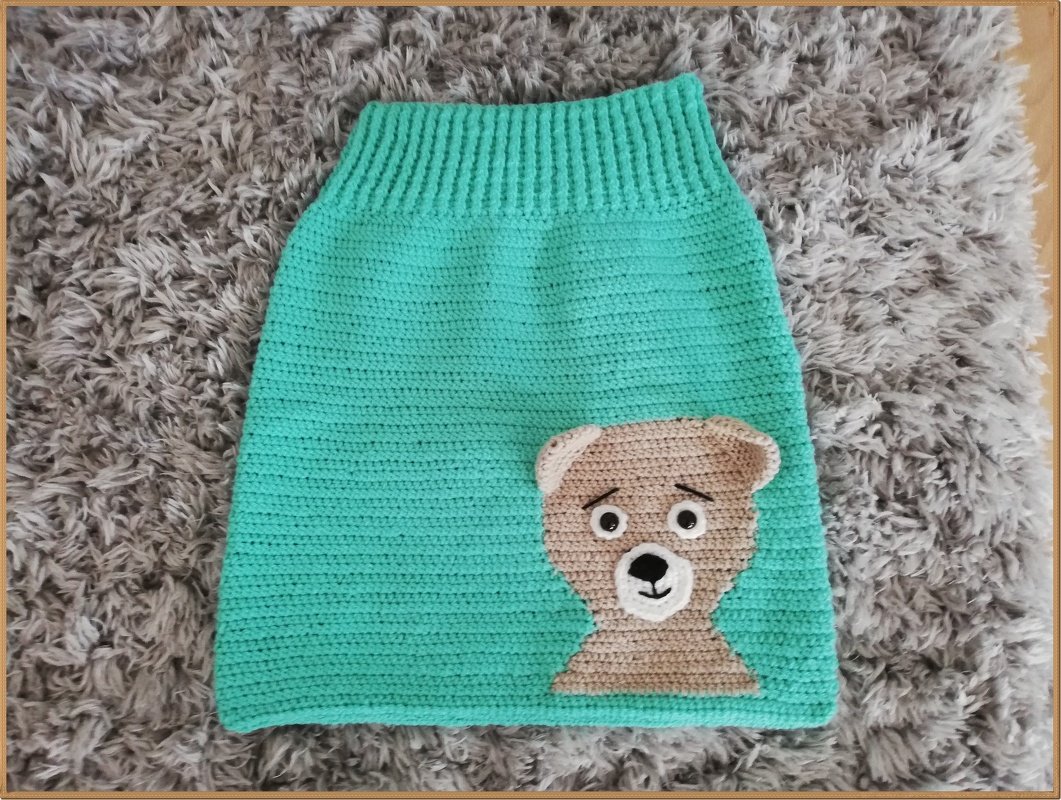 Gehäkelter türkiser Babyrock mit brauner Teddybär-Applikation auf grauem Teppich.