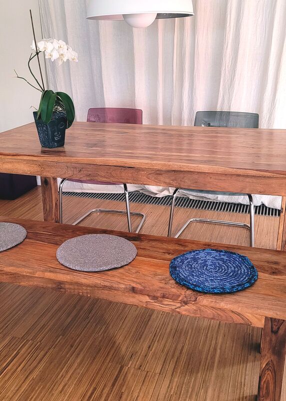 Essbereich mit Holztisch, Holzbank und drei runden Tischsets in Grau und Blau; weiße Orchidee in einem Topf mit Buddha-Motiv auf dem Tisch.