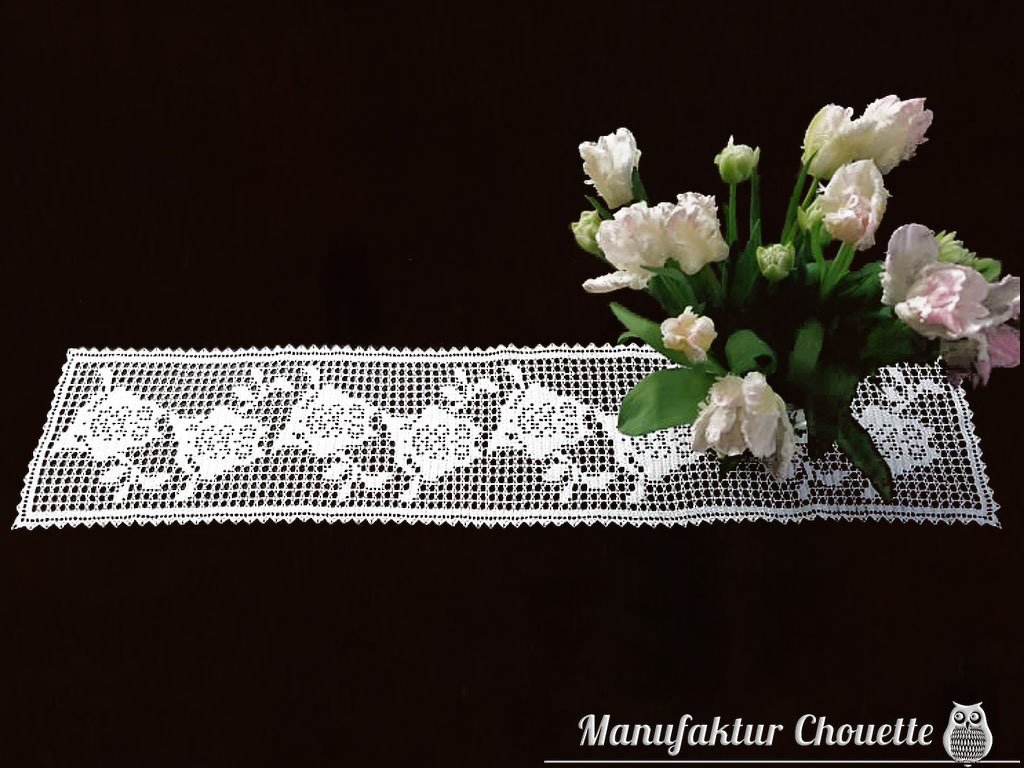 Weißer gehäkelter Tischläufer mit detailreichem Blumen-Filetmuster auf dunklem Untergrund, daneben ein heller Blumenstrauß.