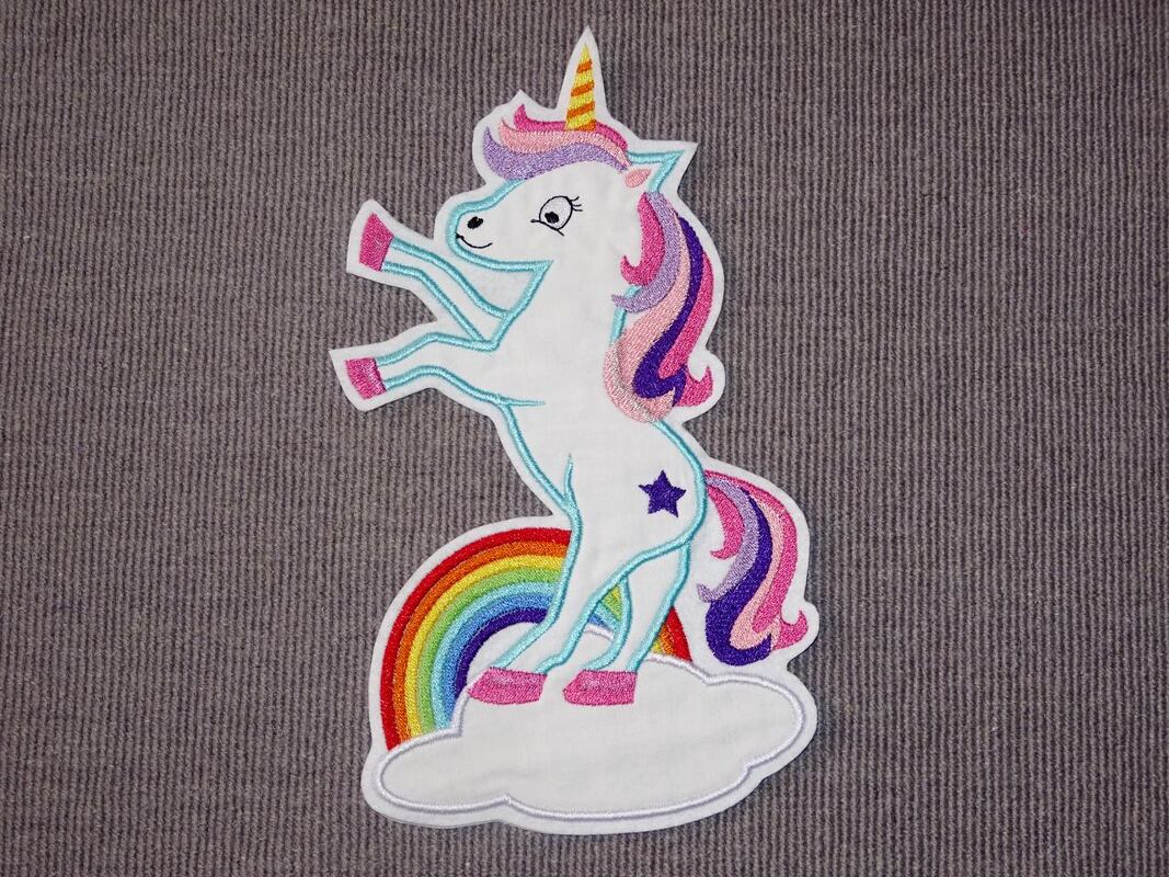 Weißes Einhorn-Stickmotiv mit rosa-violetter Mähne und Schweif, gelbem Horn, pinken Hufen und kleinem lila Stern auf der Flanke, auf einer weißen Wolke vor einem Regenbogen.