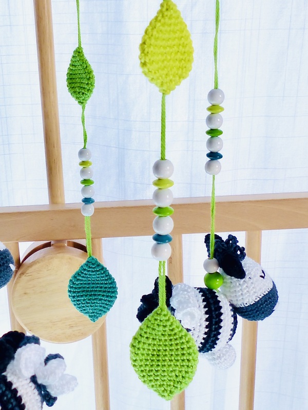 Gehäkeltes Mobile mit grünen und türkisfarbenen Blättern, weißen, grünen und türkisfarbenen Perlen und gestreiften Tieranhängern an Holzlatten eines Babybetts