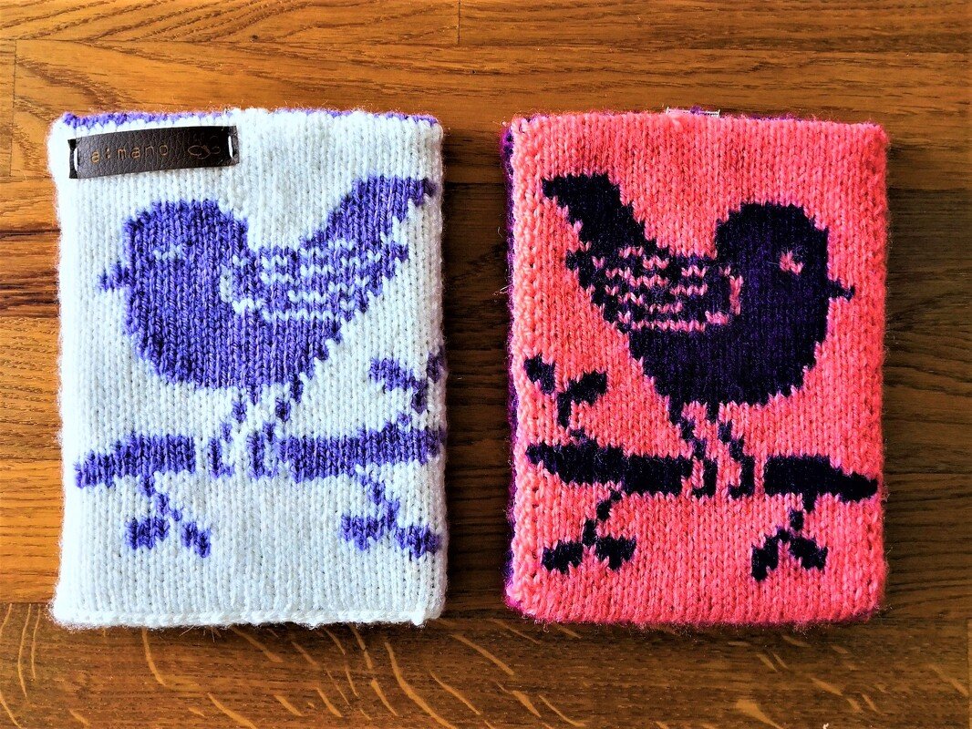 Zwei gestrickte Untersetzer auf Holz, links weiß mit lila Vogelmotiv, rechts pink mit dunkelviolettem Vogelmotiv.