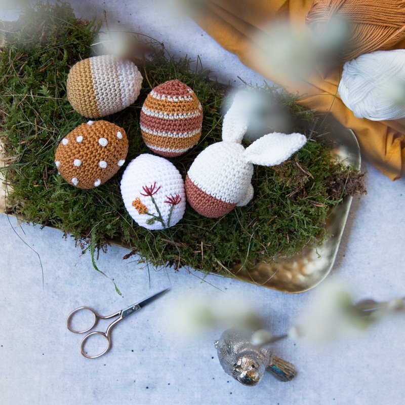 Gehäkelte Ostereier und ein Häkelhäschen auf Moos in einer goldenen Schale, daneben Schere und Wollknäuel.