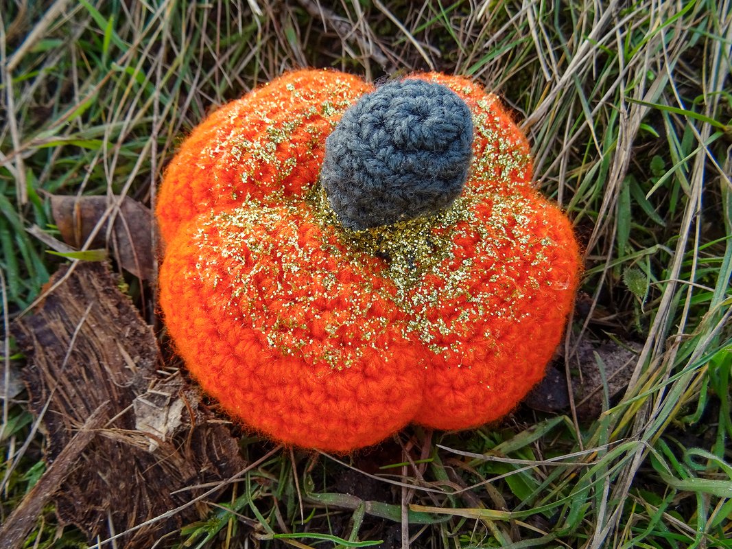 Gehäkelter oranger Kürbis mit grauem Stiel und goldenem Glitzer im Gras