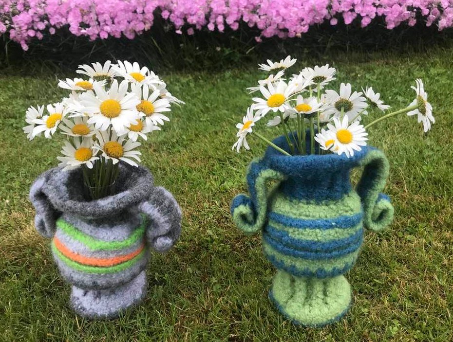 Zwei bunte gehäkelte Vasenbezüge mit Gänseblümchen auf einer Wiese vor rosa Blüten.