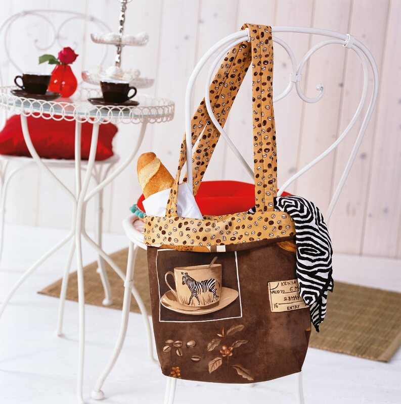 Geräumige braune Einkaufstasche aus Stoff mit Kaffeebohnenmuster, Zebra-Tassenmotiv und einem Baguette, hängt an einem weißen Stuhl.