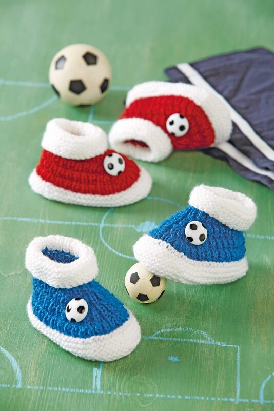 Vier gestrickte Babyschuhe in Rot und Blau mit Fußballknöpfen auf grünem Spielfeld