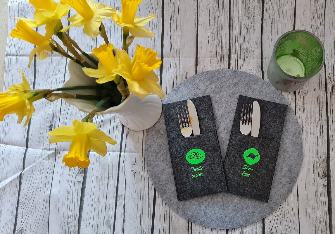 Zwei dunkelgraue Filz-Bestecktaschen mit neongrünen Schildkröten-Plotmotiven und englischen Schriftzügen, auf rundem Filzuntersetzer platziert; links weiße Vase mit gelben Narzissen und rechts ein grünes Glas