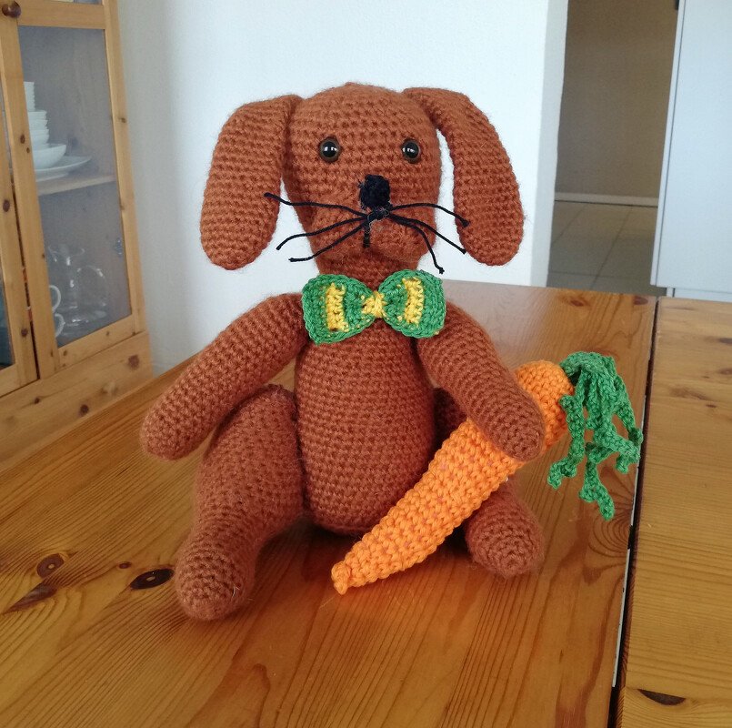 Brauner gehäkelter Amigurumi-Hase mit grüner Fliege und Karotte auf einem Holztisch