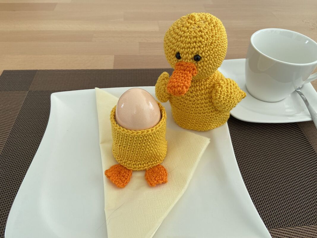 Gehäkelte gelbe Ente und Eierwärmer mit Entenfüßen auf einem weißen Teller neben einer Tasse