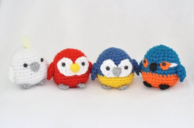 Vier kleine gehäkelte Amigurumi-Vögel nebeneinander in Weiß, Rot, Blau-Gelb und Türkis-Orange
