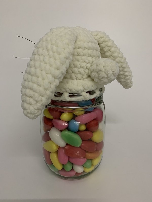 Ein Einmachglas voller pastellfarbener Dragees, bedeckt mit einer abnehmbaren weißen gehäkelten Hasenmütze mit Schlappohren