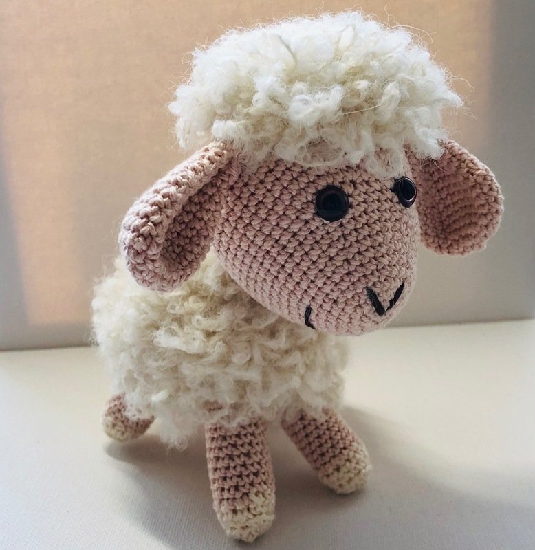 Gehäkeltes Schaf‑Amigurumi mit cremeweißem, flauschigem Körper und zartrosa Kopf, Ohren und Beinen; schwarze Augen, gestickter Mund.