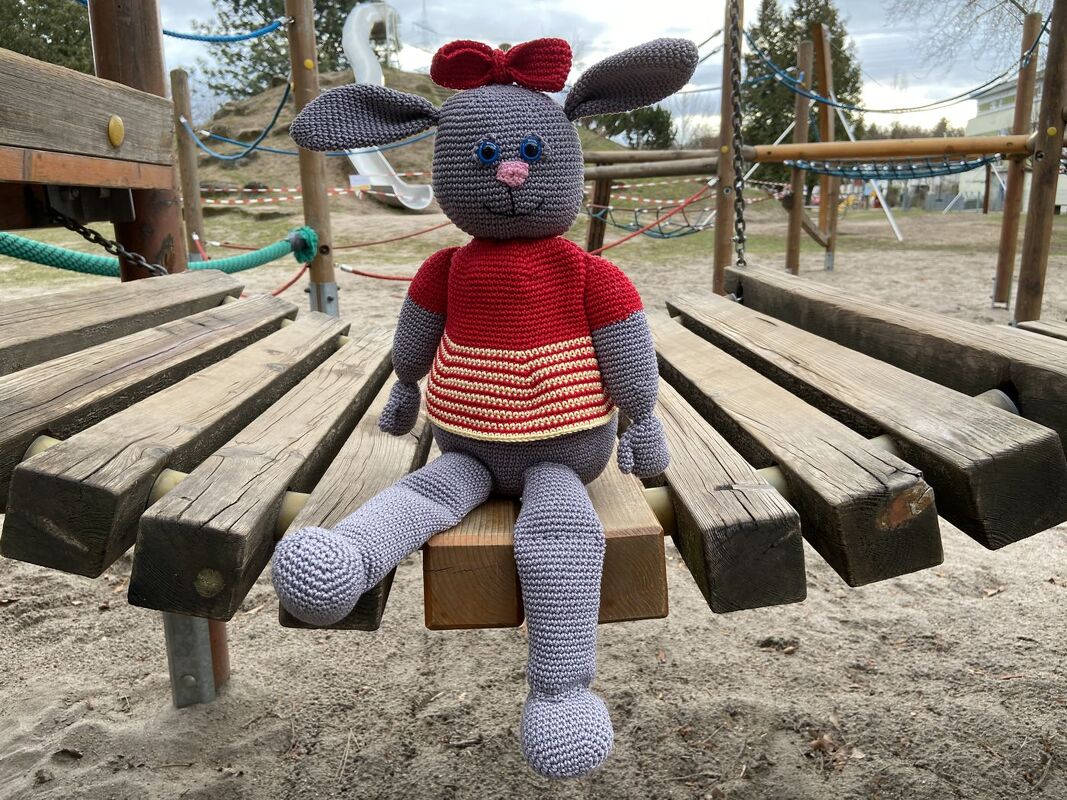 Gehäkelter grauer Amigurumi-Hase mit rotem Pullover und Schleife sitzt auf einer Holzbrücke