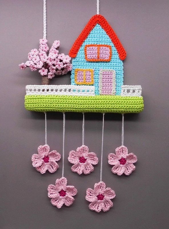 Crocheted wall hanging of a small blue house with an orange roof, pink blossom tree, white fence on a green base, and five hanging pink flowers.