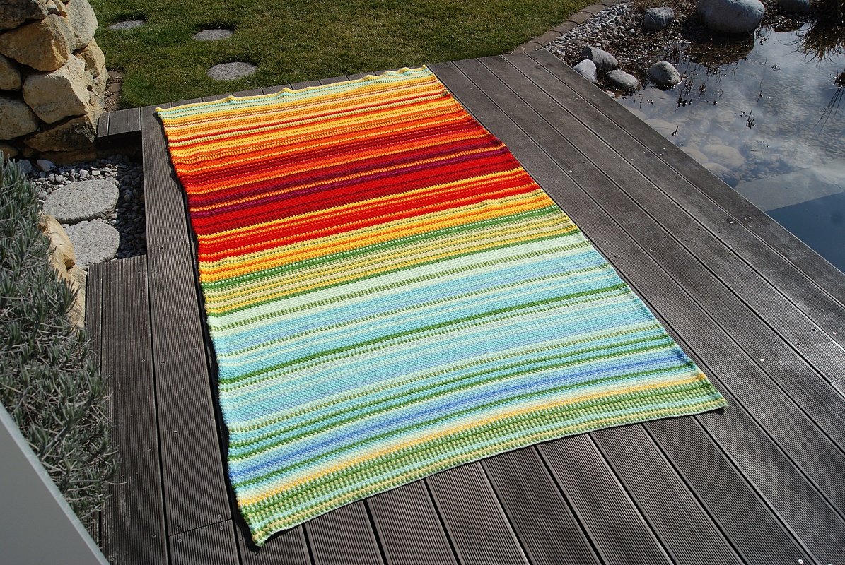 Bunte gehäkelte Decke mit warmen Rot- und Gelbtönen oben und kühlen Blau-Grün-Tönen unten auf einer Terrasse neben einem Teich