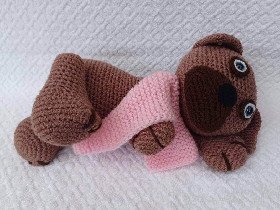 Gehäkelter brauner Amigurumi Hund mit rosa Schal auf weißem Untergrund liegend