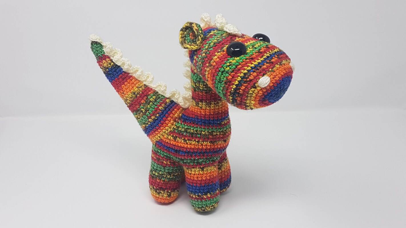Amigurumi-Dinosaurier in Regenbogenstreifen mit weißen Zacken und schwarzen Augen auf weißem Hintergrund.