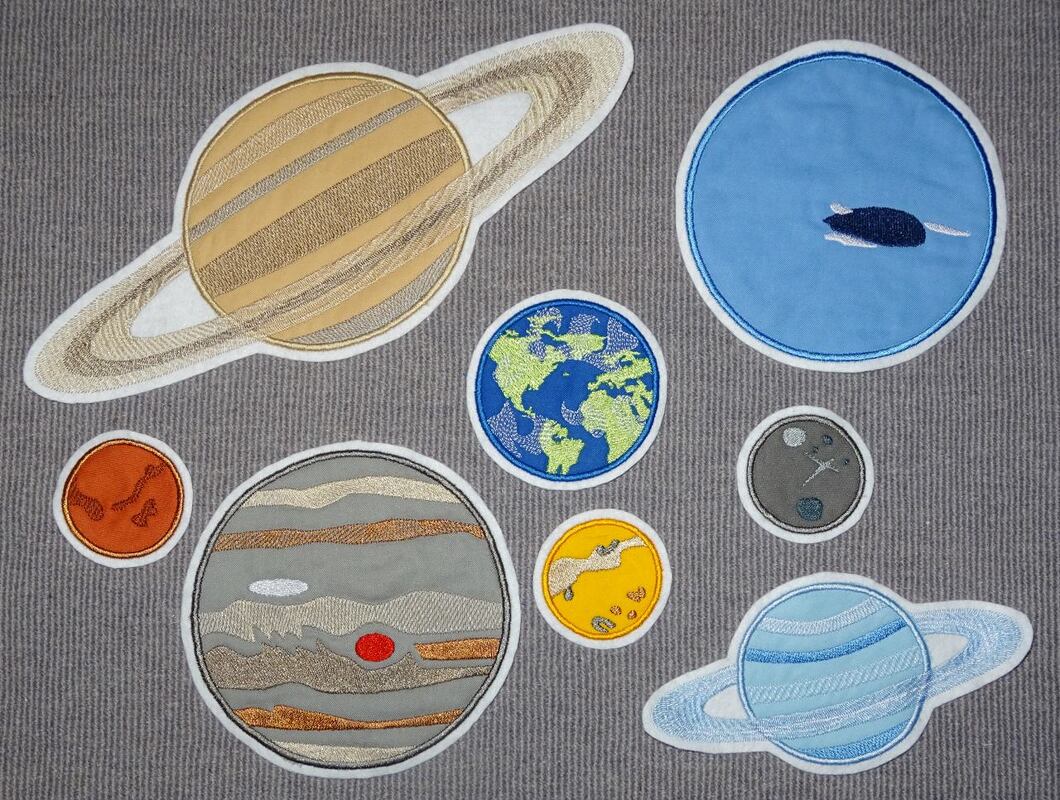 Acht bestickte Planetentextil-Patches (z. B. Saturn, Jupiter, Erde, Mars, Mond) auf grauem Stoffhintergrund.