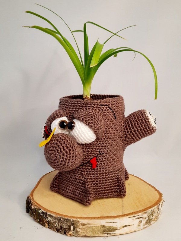 Crocheted brown plant pot cover shaped like a tree stump character with large eyes and a green plant, displayed on a wooden slab.