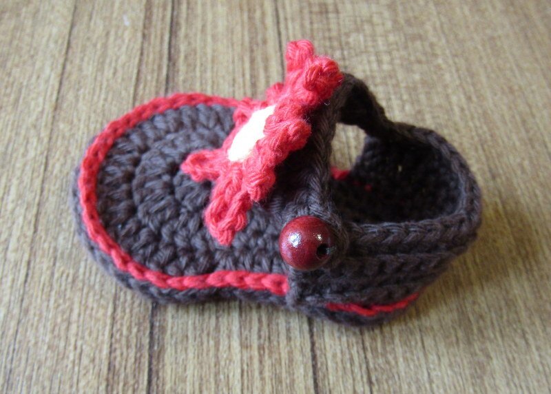Gehäkelter Babyschuh mit Riemen, rosafarbenem Zierrand, Blüte und Holzknopf auf Holzuntergrund