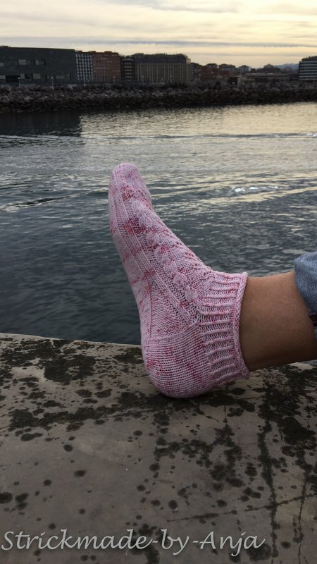 Fuß mit rosa-melierter handgestrickter Knöchelsocke vor Wasser