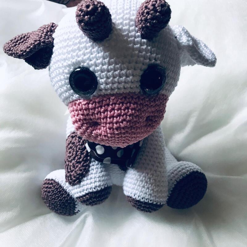 Gehäkelte Amigurumi-Kuh in Weiß und Braun mit rosa Schnauze und gepunktetem Halsband.