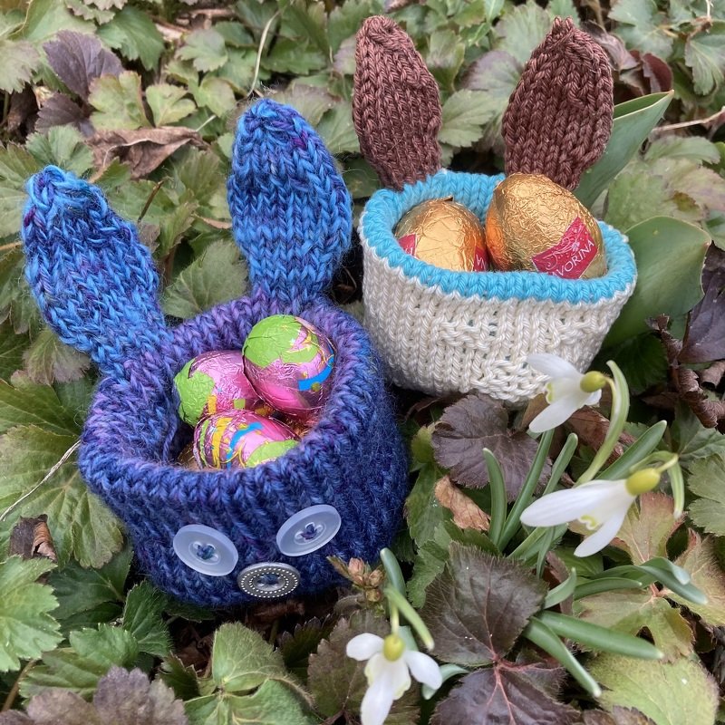 Zwei gestrickte Körbchen in Hasenform in Blau und Natur, mit Ohren und Knopfaugen, gefüllt mit Schokoeiern zwischen Blättern und Schneeglöckchen