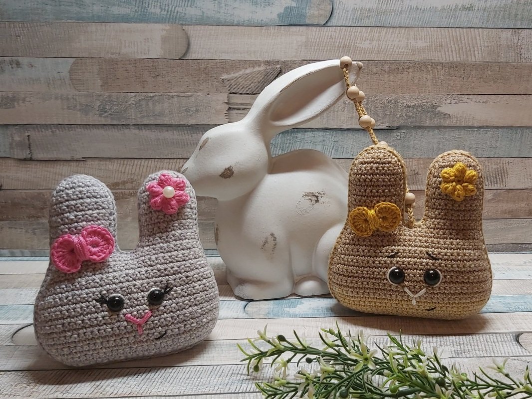 Zwei gehäkelte Hasenfiguren, grau mit pinker Blume und beige mit gelber Blume, vor einer weißen Hasenfigur auf Holzlatten