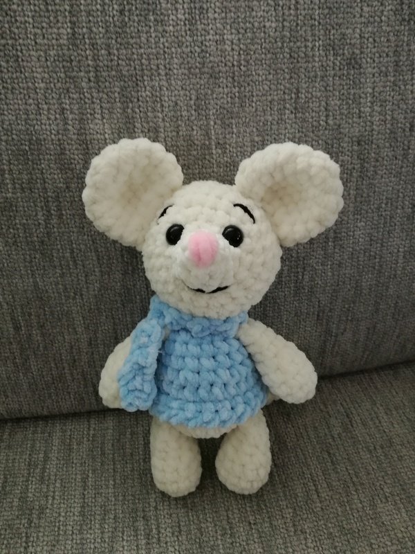 Gehäkelte weiße Amigurumi-Maus mit hellblauem Pulli auf grauem Sofa