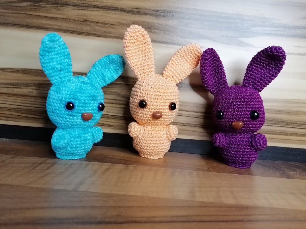 Drei gehäkelte Amigurumi-Hasen in Türkis, Pfirsich und Violett auf einem Holzboden