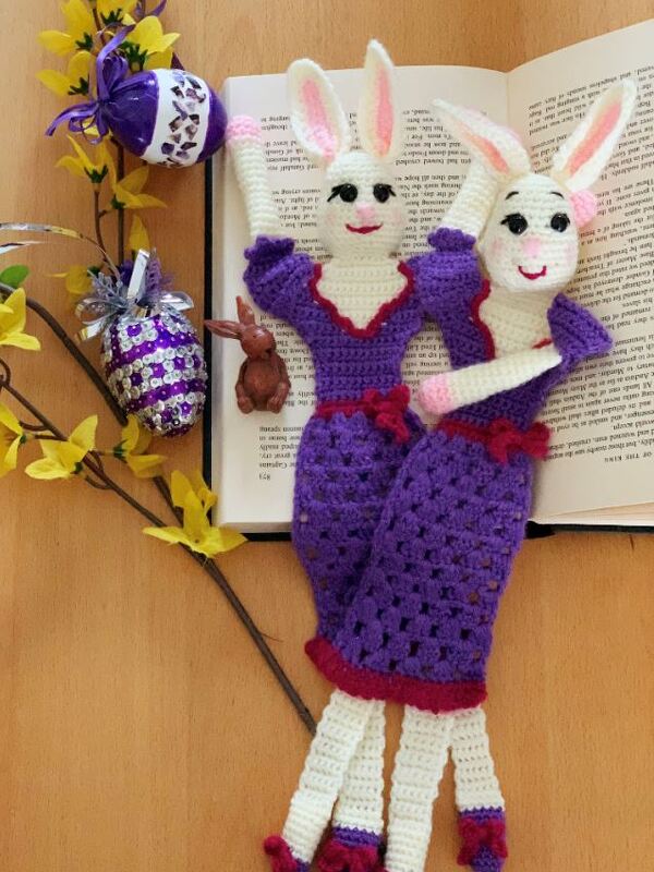 Zwei gehäkelte Hasen-Lesezeichen in Lila und Weiß auf einem aufgeschlagenen Buch, umgeben von gelben Blüten und dekorativen Ostereiern.