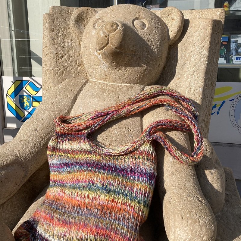 Multicolored knitted shoulder bag with long strap draped on a carved stone bear bench