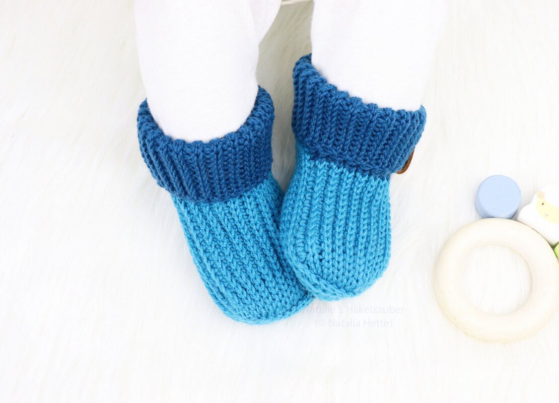 Zwei blaue Babyschuhe mit geripptem Umschlagbündchen auf weißem, flauschigem Untergrund.