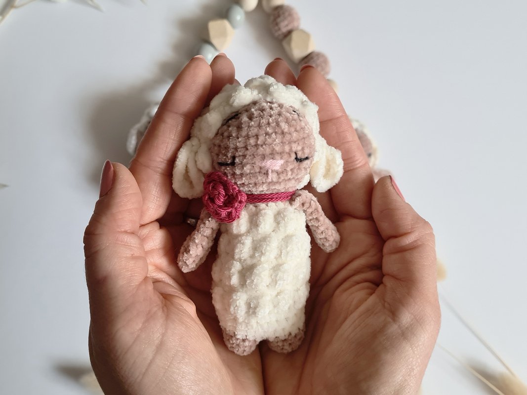 Gehäkeltes Mini-Lämmchen mit weißem flauschigem Körper und rosa Blüte, in Händen gehalten