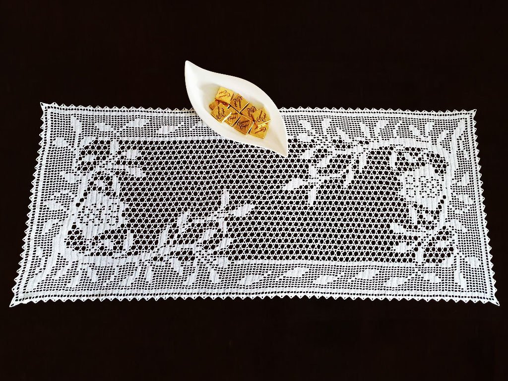 Weißer rechteckiger Spitzentischläufer mit filigranem Häkelmuster und einer kleinen Schale mit Pralinen auf dunklem Untergrund