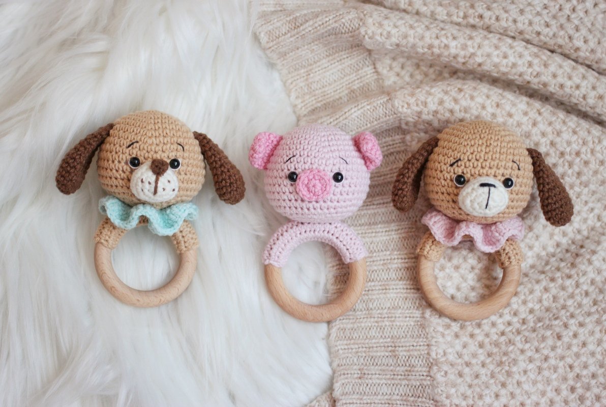 Three crochet baby rattles: two brown dogs with blue and pink ruffles, and one pink pig, all with wooden rings, on a fluffy white and knitted beige background.