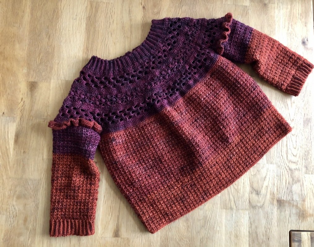 Gehäkelter Kinderpullover in Dunkelviolett und Rostrot mit Lochmuster am Oberteil und Rüschen an den Ärmeln, liegend auf einem Holztisch.