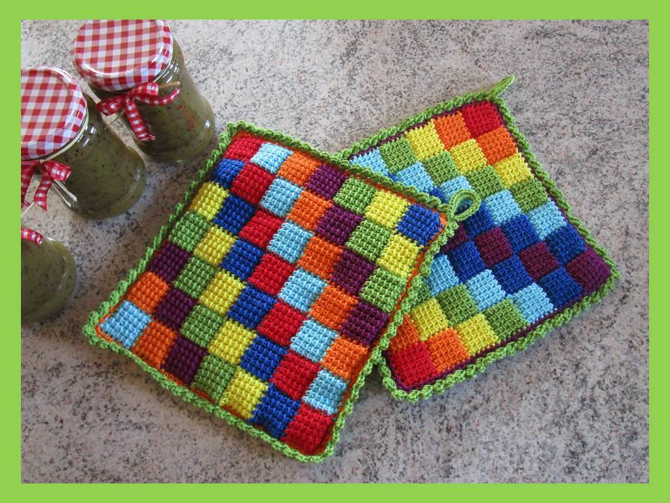 Zwei bunte handgehäkelte Topflappen mit kariertem Muster und grüner Umrandung auf einer Arbeitsfläche; im Hintergrund drei Gläser mit karierten Deckeln.