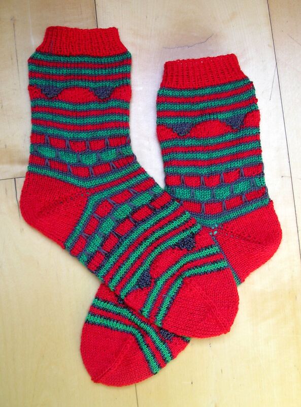 Paar gestrickter Damensocken in Rot und Grün mit Streifen, Herz- und Quadratmustern auf hellem Holzuntergrund.