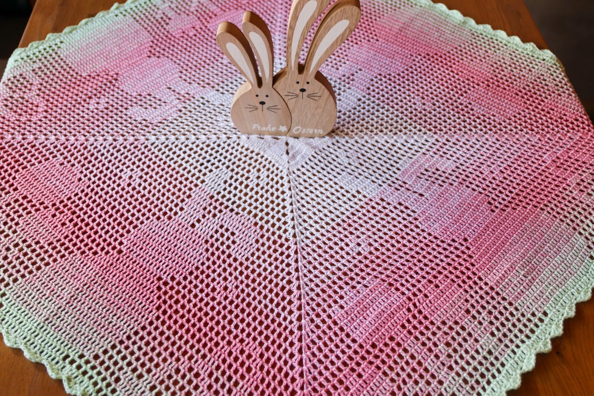 Circular ombre crochet doily in pink-to-green with openwork pattern on a wooden table and two wooden bunny figurines.